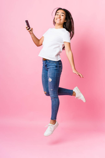 Young indian woman holding phone jumping on pink background - Foto, Imagem