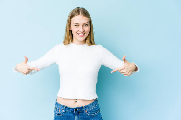 Young blonde woman isolated on blue background points down with fingers, positive feeling. - Photo, Image