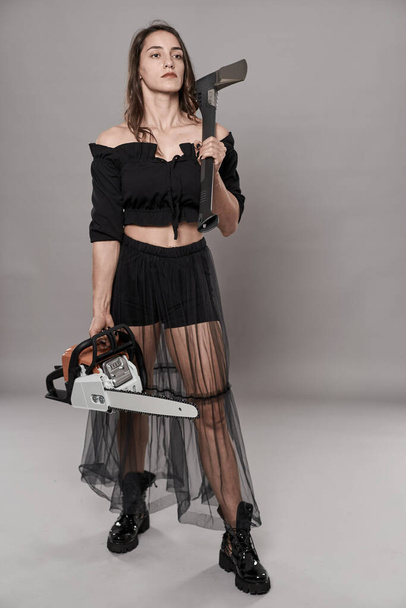 Female villain holding a chainsaw and an axe, with sobering expression - Photo, Image