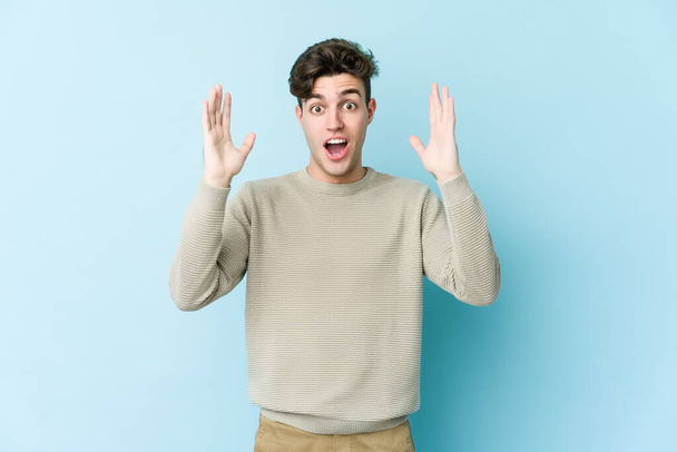 Young caucasian man isolated on blue background celebrating a victory or success, he is surprised and shocked. - Foto, Imagen