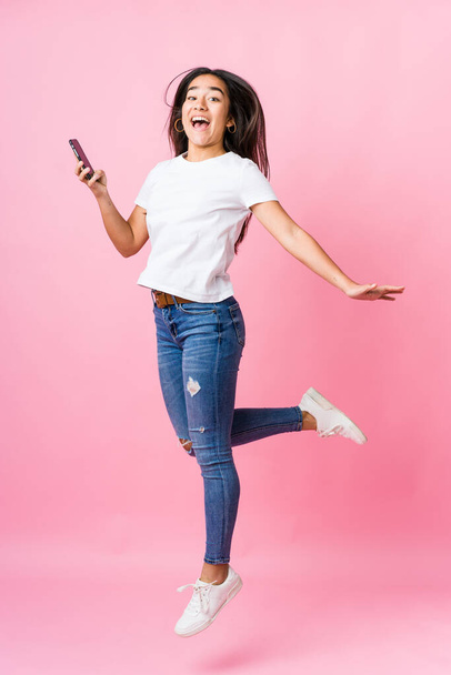 Young indian woman holding phone jumping on pink background - Photo, Image