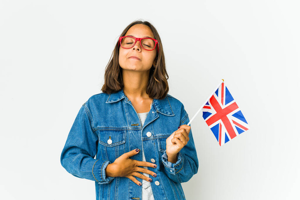 Young latin woman holding a english flag isolated on white background touches tummy, smiles gently, eating and satisfaction concept. - Photo, Image