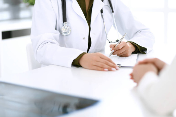 Woman doctor and patient sitting and talking at medical examination at hospital office, close-up. Therapist filling up medication history records. Medicine and healthcare concept - Foto, imagen