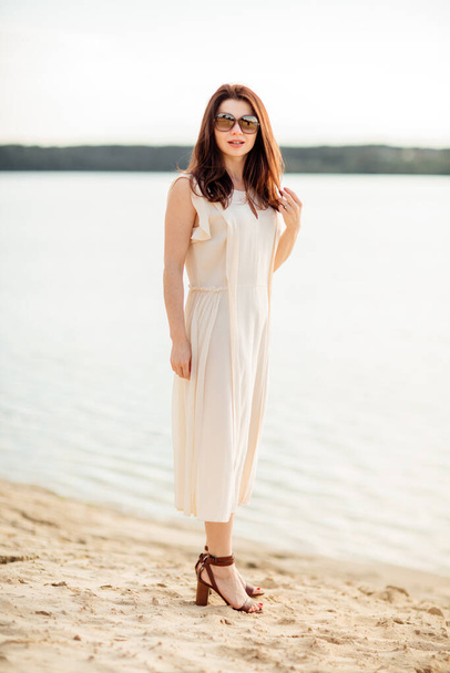 Jolie jeune femme aux cheveux foncés près de la plage près du lac en robe et lunettes de soleil. concept de vacances heureuses et temps de villégiature. - Photo, image