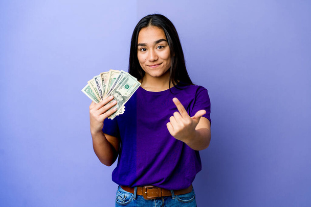 jonge aziatische vrouw houden geld geïsoleerd op paarse achtergrond wijzend met de vinger naar u alsof uitnodigen dichterbij komen. - Foto, afbeelding