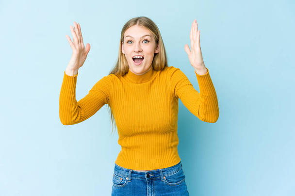 Νεαρή ξανθιά γυναίκα απομονωμένη σε μπλε φόντο λαμβάνει μια ευχάριστη έκπληξη, ενθουσιασμένοι και σηκώνοντας τα χέρια. - Φωτογραφία, εικόνα