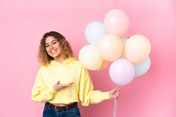 Young blonde woman with curly hair catching many balloons isolated on pink background extending hands to the side for inviting to come - Foto, Imagem