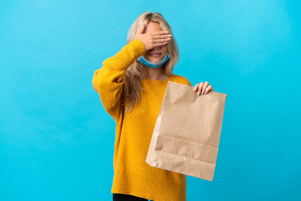 Молодая россиянка, держащая сумку с продуктами на синем фоне, закрывает руками глаза. Не хочу ничего видеть. - Фото, изображение