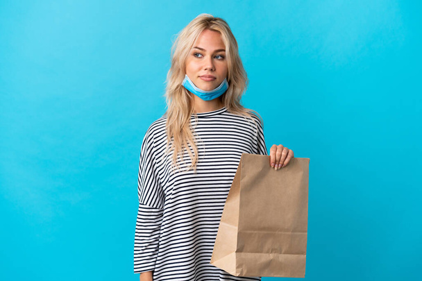 Joven mujer rusa sosteniendo una bolsa de la compra de comestibles aislada sobre fondo azul que tiene dudas mientras mira al lado - Foto, Imagen
