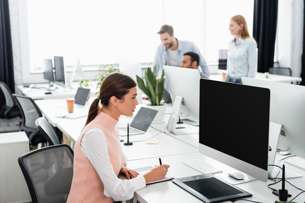 Бізнес-леді пишуть на ноутбуці біля комп'ютера, графічного планшета та багатоетнічних колег на розмитому фоні в офісі
  - Фото, зображення