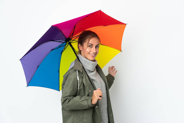 Mujer joven sosteniendo un paraguas aislado sobre fondo blanco extendiendo las manos a un lado para invitar a venir - Foto, Imagen