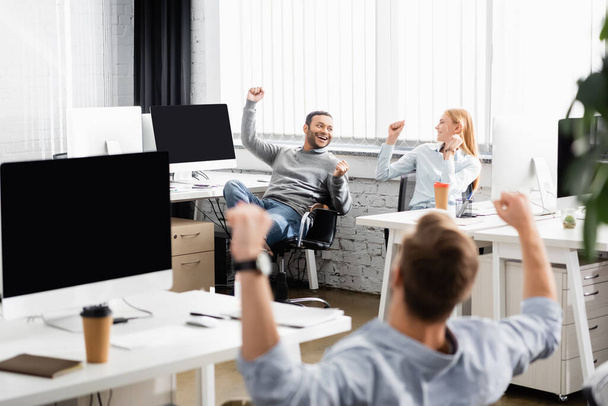Multiethnic businesspeople showing yeah gesture while working near computers in office  - Foto, Bild