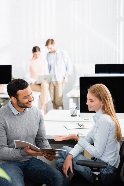 Lächelnde multiethnische Geschäftsleute blicken auf Papiermappe, während Kollegen im Büro an verschwommenem Hintergrund arbeiten  - Foto, Bild