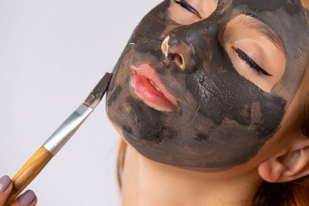 Enlarged photo of a young woman with closed eyes applying a clay mask to her face with a brush. Photo on white background. High quality photo - Foto, Imagen