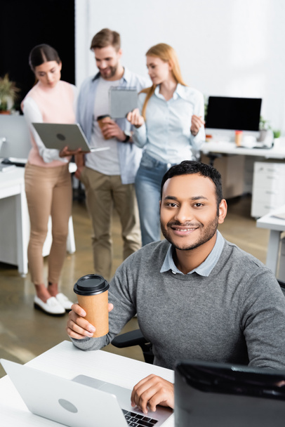 Sorrindo índio empresário segurando café para ir ao usar laptop perto de colegas em fundo turvo  - Foto, Imagem
