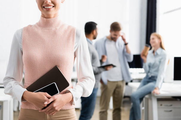 Oříznutý pohled usměvavé podnikatelky držící smartphone a notebook s kolegy na rozmazaném pozadí  - Fotografie, Obrázek