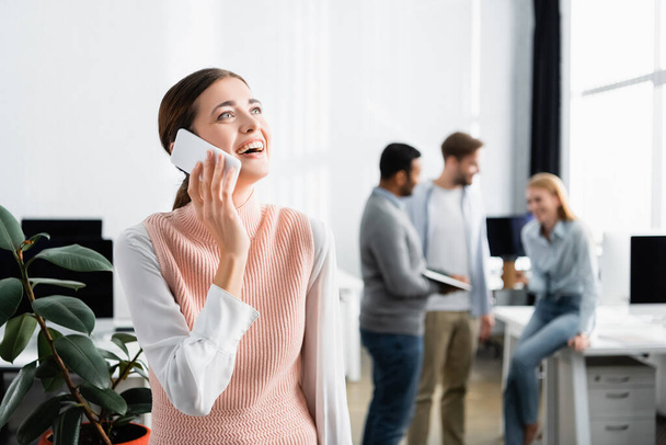 Empresária sorridente conversando no smartphone perto de colegas no fundo borrado no escritório  - Foto, Imagem