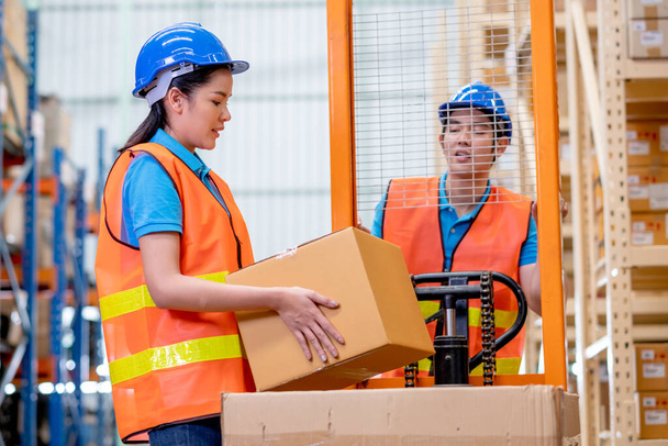 Warehouse woman or factory worker hold package box in front of hand stacker or hydraulic cart that control by her co-worker man in workplace area for delivery industrial business. - Fotó, kép