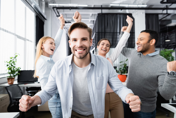 Smiling businessman showing yeah gesture near cheerful multiethnic colleagues on blurred background in office  - Foto, imagen