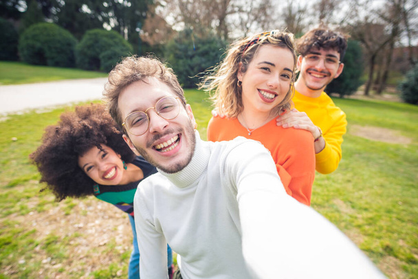 Група з чотирьох багатоетнічних різноманітних друзів на відкритому повітрі за допомогою смартфона, що приймає селфі, розважаючись разом у парку, посміхаючись
  - Фото, зображення
