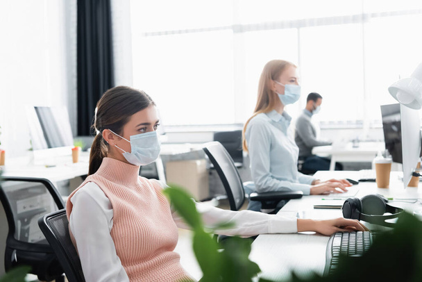 Businesswoman in medical mask sitting near computer keyboard and colleagues on blurred background  - Fotoğraf, Görsel