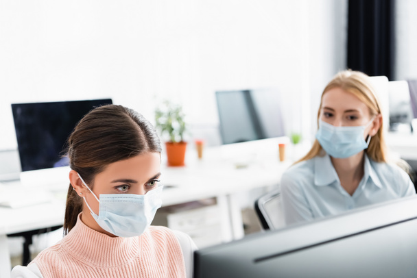 Businesswoman in medical mask looking at computer monitor near colleague in office  - 写真・画像