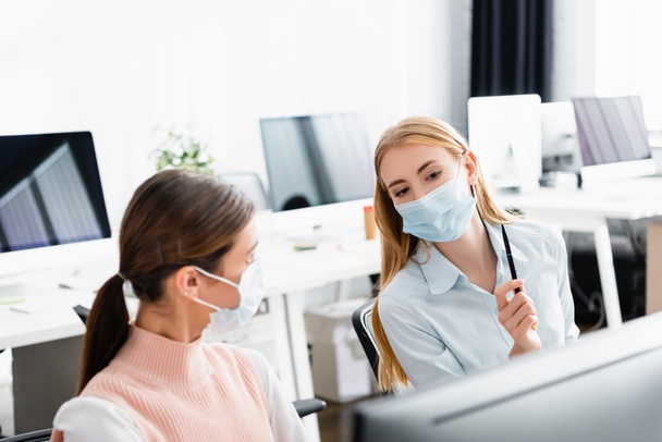 Businesswomen in medical masks talking near computer in office  - Valokuva, kuva