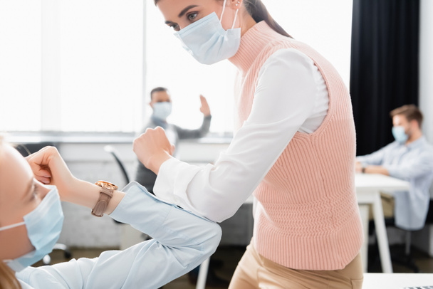 Businesswomen in medical masks giving high five with colleagues on blurred background in office  - Photo, Image