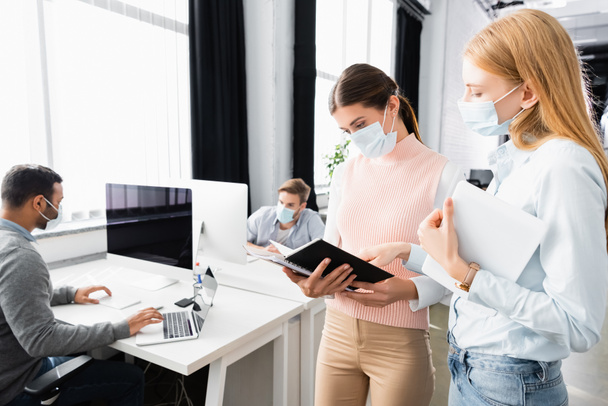 Businesswomen in medical masks with digital table looking at notebook near colleagues on blurred background  - Foto, Imagem