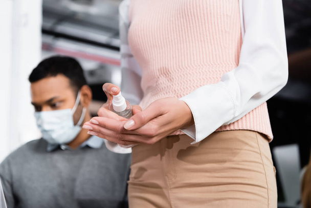 Vue recadrée de femme d'affaires en utilisant un désinfectant pour les mains près d'un collègue indien dans un masque médical sur fond flou  - Photo, image