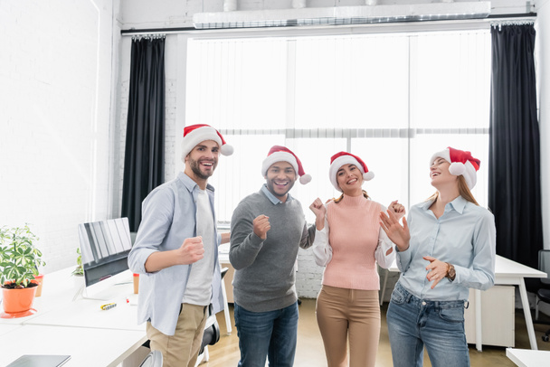 Cheerful multiethnic businesspeople in santa hats showing yeah gesture in office  - Foto, Imagem