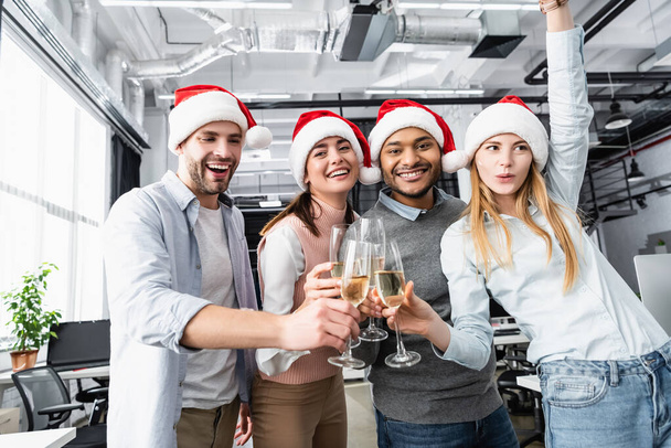 Positive multiethnic businesspeople in santa hats clinking with champagne in office  - Fotografie, Obrázek