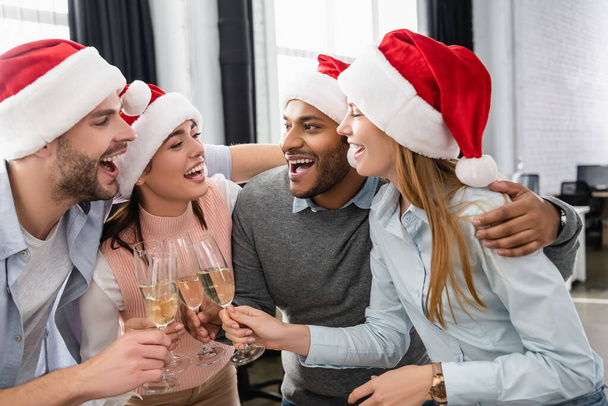 Multiethnic businesspeople in santa hats embracing while clinking with champagne in office  - Foto, immagini