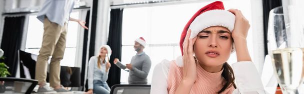 Drunk businesswoman in santa hat sitting near glass of champagne and colleagues on blurred background in office, banner  - Valokuva, kuva