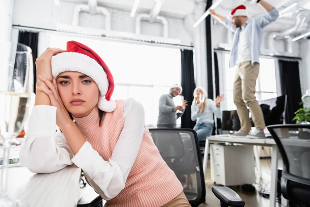 Tired businesswoman in santa hat looking at camera near glass of champagne and colleagues on blurred background in office  - Zdjęcie, obraz
