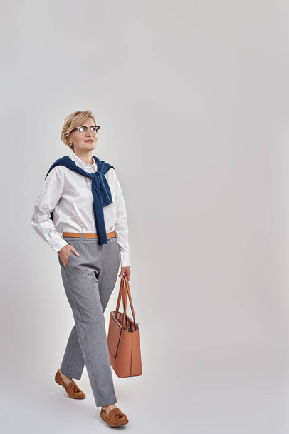 Full length shot of elegant middle aged caucasian woman wearing business attire and glasses looking away while posing with handbag isolated over grey background - Foto, afbeelding