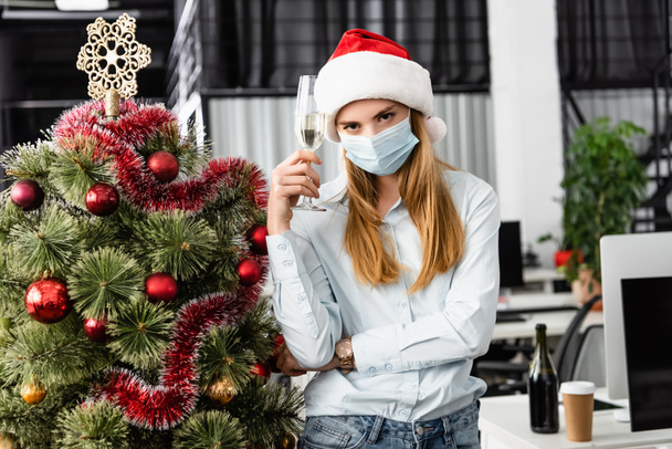 Бизнесмен в медицинской маске держит бокал шампанского возле рождественской елки в офисе  - Фото, изображение