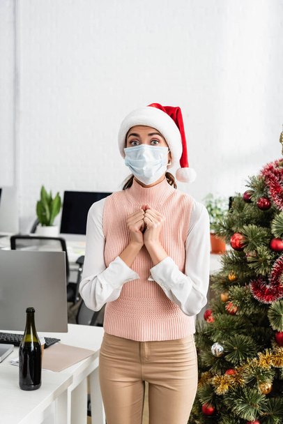Jeune femme d'affaires en chapeau de Père Noël et masque médical debout près de l'arbre de Noël et bouteille de champagne au bureau  - Photo, image