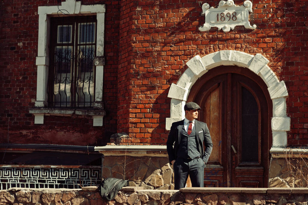 Portrait of retro 1920s english arabian business man wearing dark suit, tie and flat cap standing against old brick house 1898 year. - Foto, afbeelding