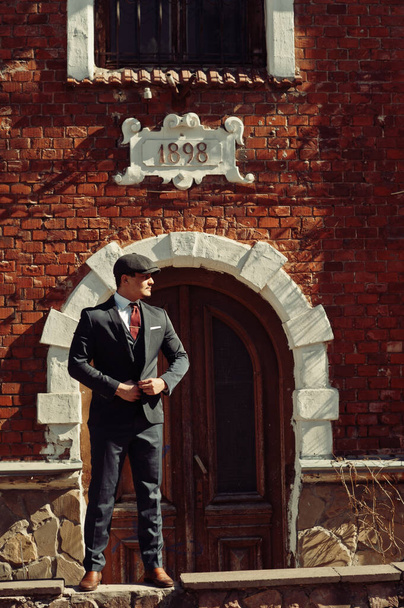 Portrait of retro 1920s english arabian business man wearing dark suit, tie and flat cap standing against old brick house 1898 year. - Zdjęcie, obraz