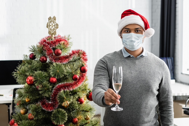 Индийский бизнесмен в медицинской маске и шляпе Санты держит бокал шампанского возле рождественской елки в офисе  - Фото, изображение