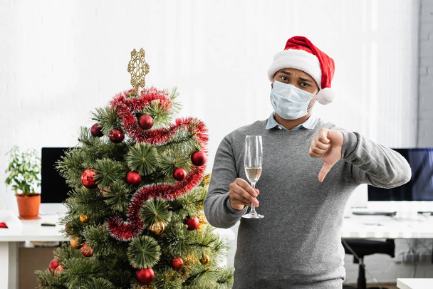 Индийский бизнесмен в медицинской маске держит бокал с шампанским и демонстрирует неприязненный жест возле рождественской елки в офисе  - Фото, изображение