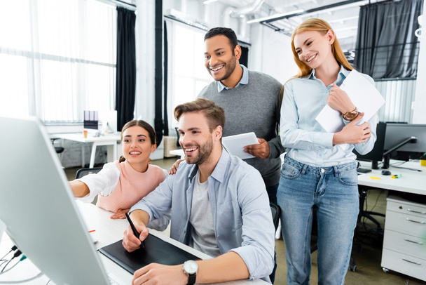 Веселые мультиэтнические предприниматели, стоящие рядом с коллегами с помощью компьютера и графического планшета в офисе  - Фото, изображение