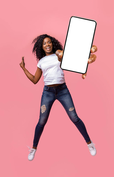 ジャンプしながら空のスマートフォンの画面を示す幸せなアフリカの女性 - 写真・画像