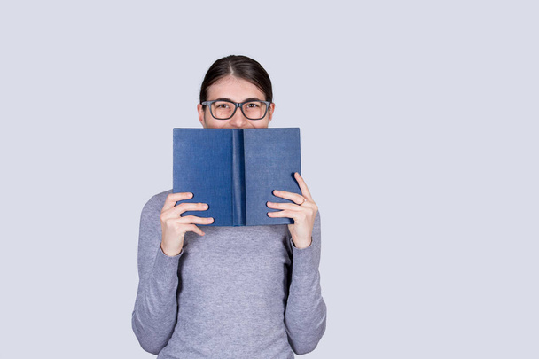 Felice studentessa con gli occhiali sorridente che copre mezza faccia con un libro blu aperto. Amante del libro. Pronti a studiare sodo. - Foto, immagini