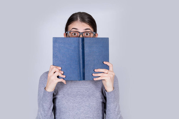 Zaskoczona uczennica w okularach zasłaniająca pół twarzy otwartą niebieską książką. Oszołomiona emocjonalnie młoda kobieta wyglądająca zdumiewająco przykrywająca pół twarzy książką. - Zdjęcie, obraz