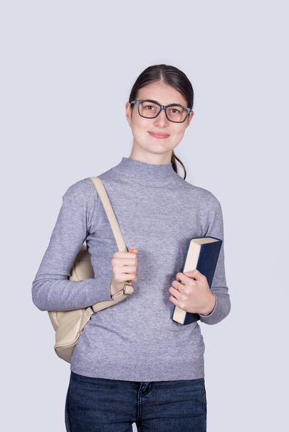 Pewna siebie studentka trzymająca jedną książkę i niosąca plecak. Wesoła uczennica z pozytywnym wyrazem twarzy odizolowana na białym tle. - Zdjęcie, obraz