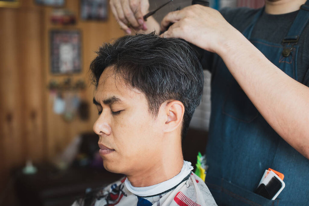 Asijský mladý muž je sestřih s nůžkami od profesionálního holiče v holičství. Kadeřník pomocí hřeben a nůžky stříhat vlasy. - Fotografie, Obrázek