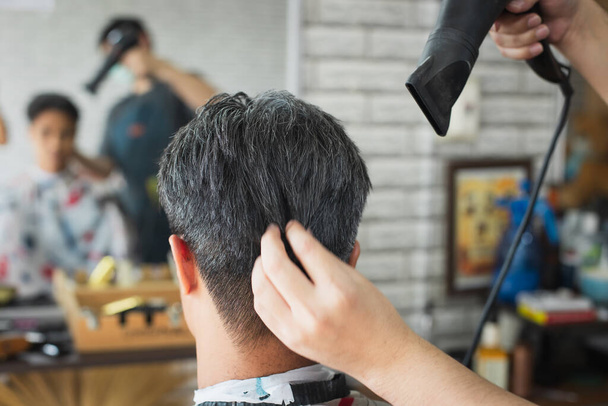 Cabeleireiro golpe secando seu cabelo cliente após o corte de cabelo acabado. ásia jovem sendo golpe seco cabelo com secador de cabelo por profissional barbeiro no barbearia. - Foto, Imagem