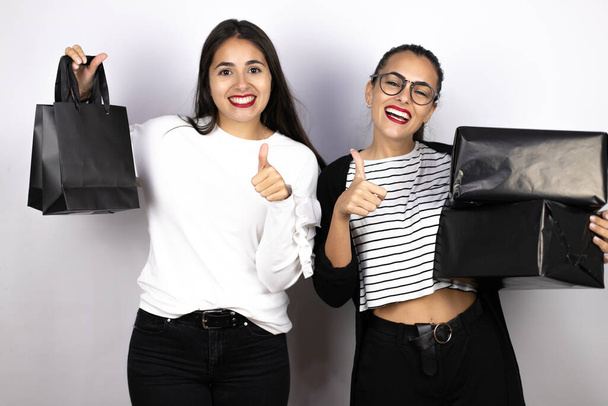 dos mujeres hermosas jóvenes sosteniendo bolsas y cajas negras haciendo la señal correcta. Viernes Negro - Foto, imagen